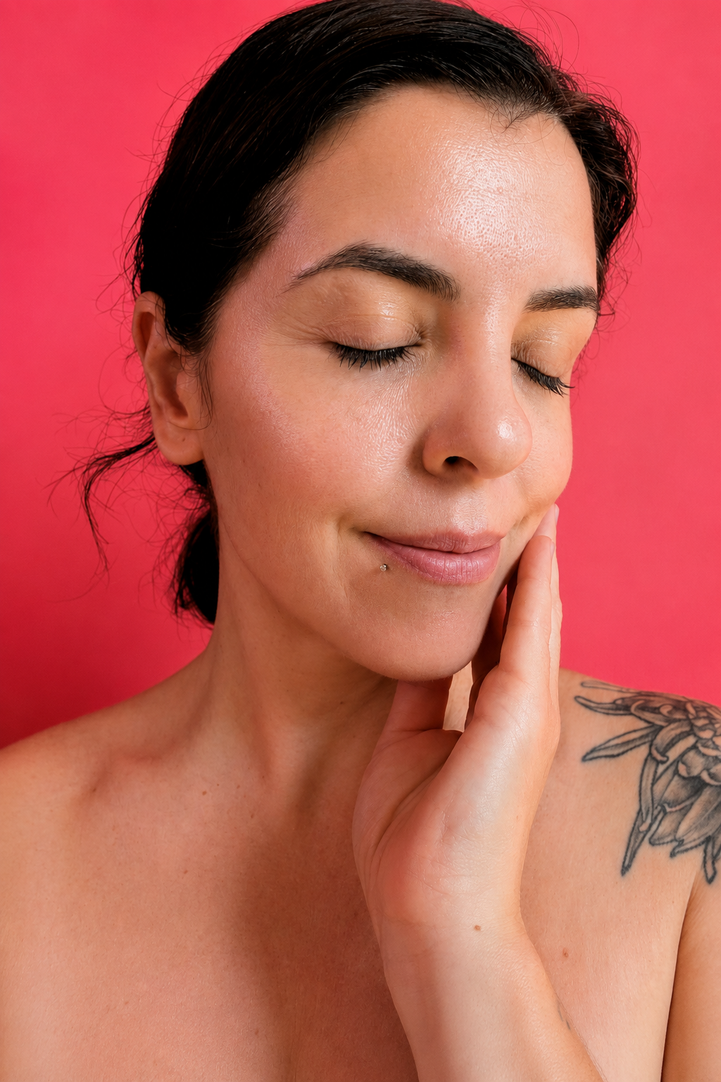 A close-up portrait of a woman with her eyes closed, touching her cheek, set against a solid pink background.